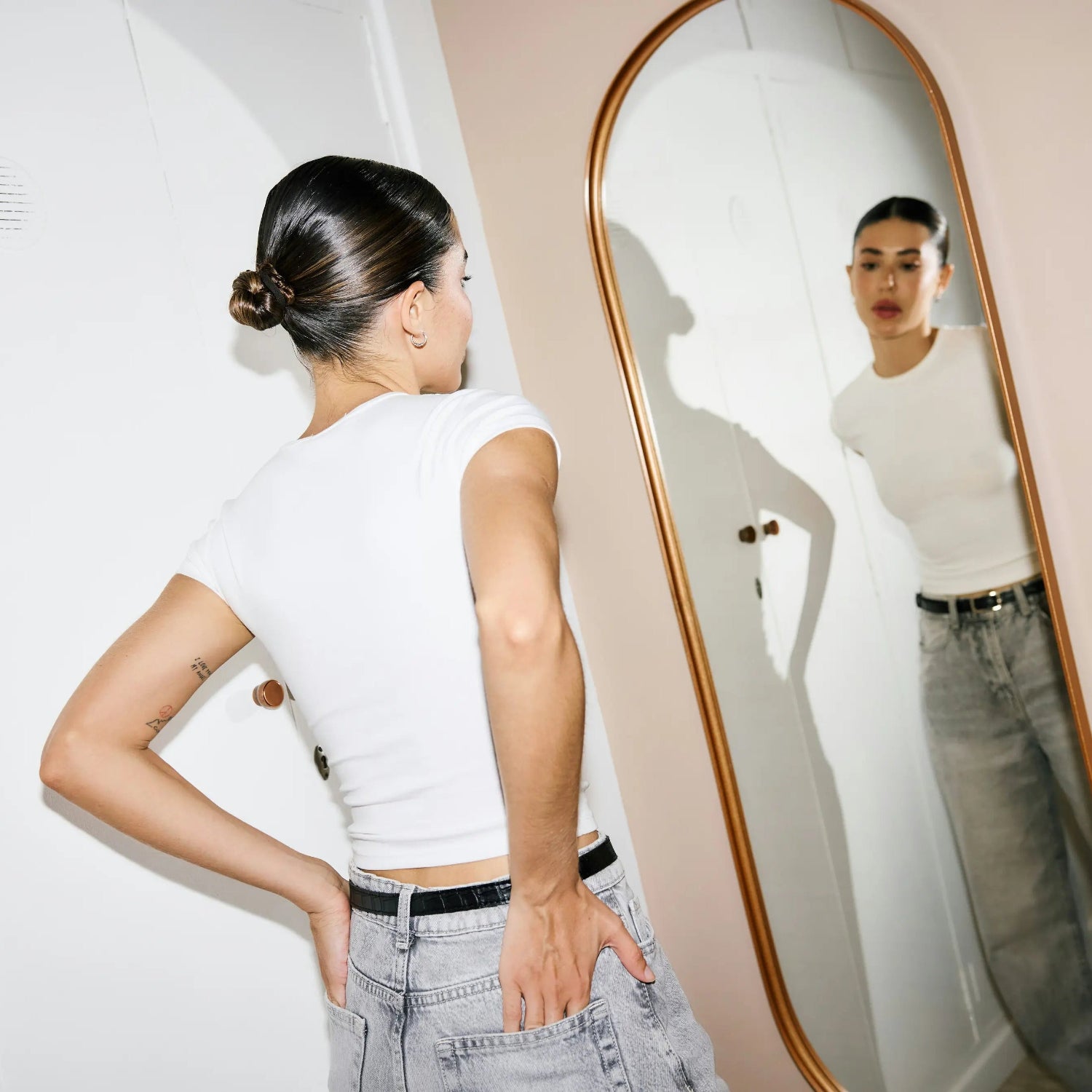 Frau betrachtet sich in einem eleganten, rundbogigen Standspiegel von Spiegelmax mit warmem Metallrahmen. Der Spiegel hängt an einer hellen Wand und reflektiert die Frau in lässigem Outfit aus weißem Shirt und Jeans.