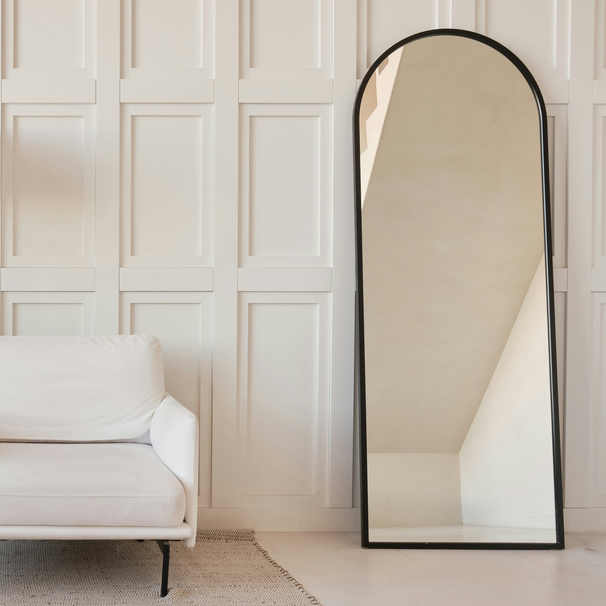 Ein eleganter, großer Standspiegel von Spiegelmax mit rundem Bogendesign und schlankem schwarzem Rahmen steht vor einer weißen Wand mit klassischer Paneeloptik. Links daneben befindet sich ein weißes Sofa, der Raum wirkt hell, modern und minimalistisch. Der Spiegel zeigt die hochwertige Verarbeitung und das klare Design, wie es für maßgefertigte Spiegel von Spiegelmax typisch ist.