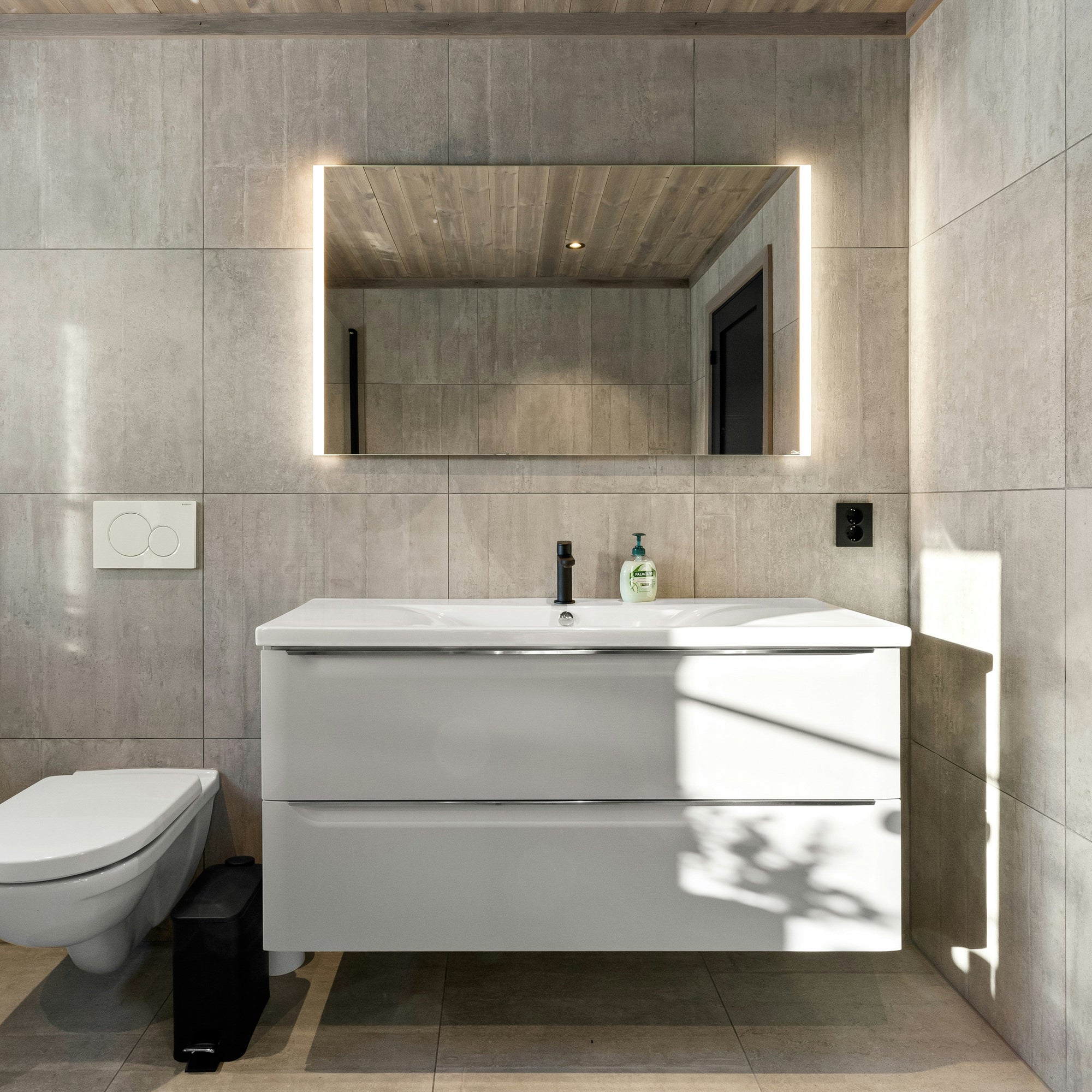 Modernes Badezimmer mit rechteckigem LED-Badspiegel von Spiegelmax, der warm hinterleuchtet ist und über einem weißen, schwebenden Waschtisch montiert wurde. Die Wände sind in grauer Betonoptik gehalten, rechts fällt Sonnenlicht ins Bad, links befindet sich ein wandhängendes WC. Die klare, reduzierte Gestaltung sorgt für ein zeitloses, minimalistisches Ambiente.