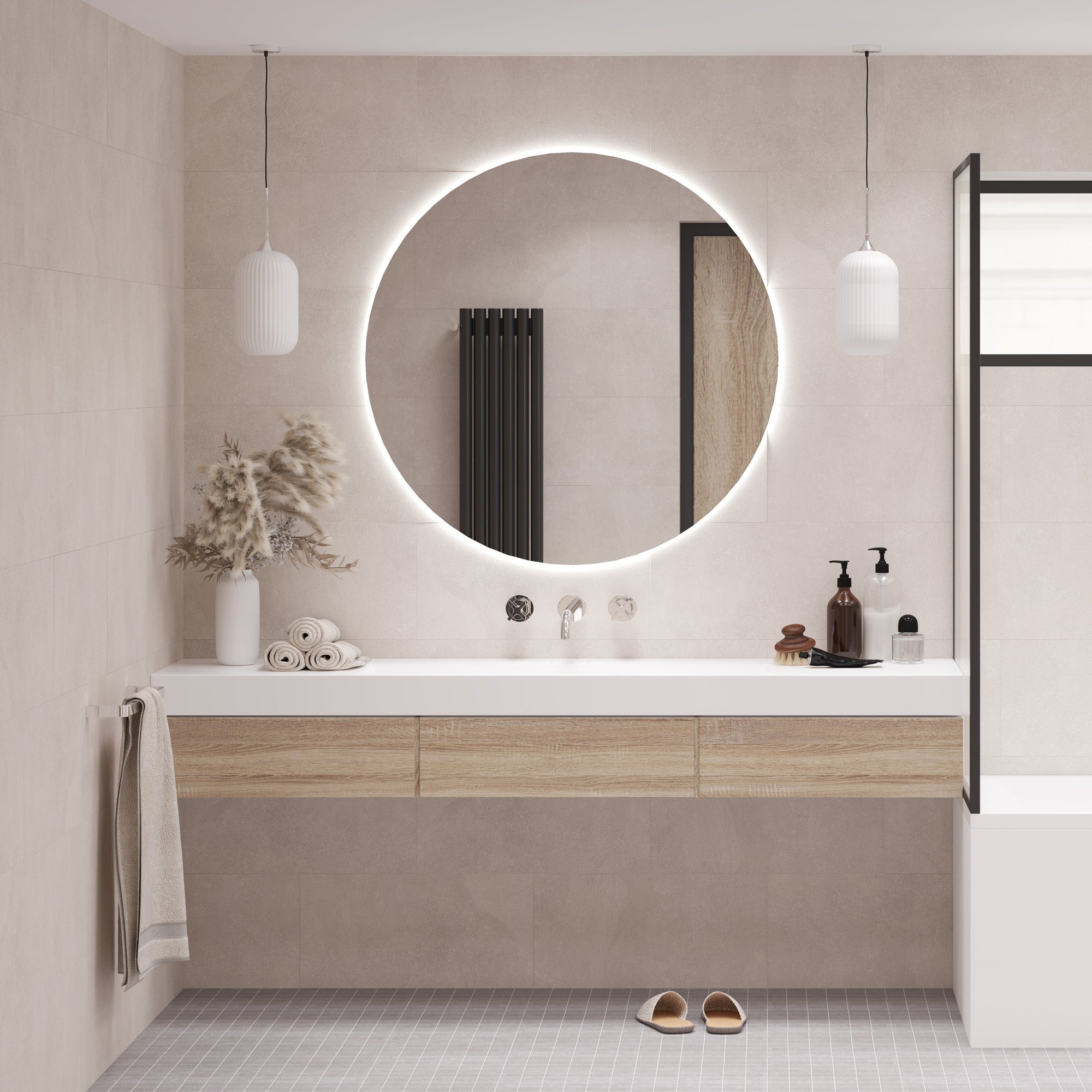 Ein großer runder LED-Badspiegel von Spiegelmax mit sanfter, umlaufender Beleuchtung hängt über einem hellen, modernen Waschtisch mit Holzakzenten. Links stehen zusammengerollte Handtücher und eine Vase mit Pampasgras, rechts befinden sich Pflegeprodukte. Zwei minimalistische Pendelleuchten rahmen den Spiegel ein, während dezente Wandfliesen und ein graues Bodenmuster ein ruhiges, elegantes Badezimmerambiente schaffen.