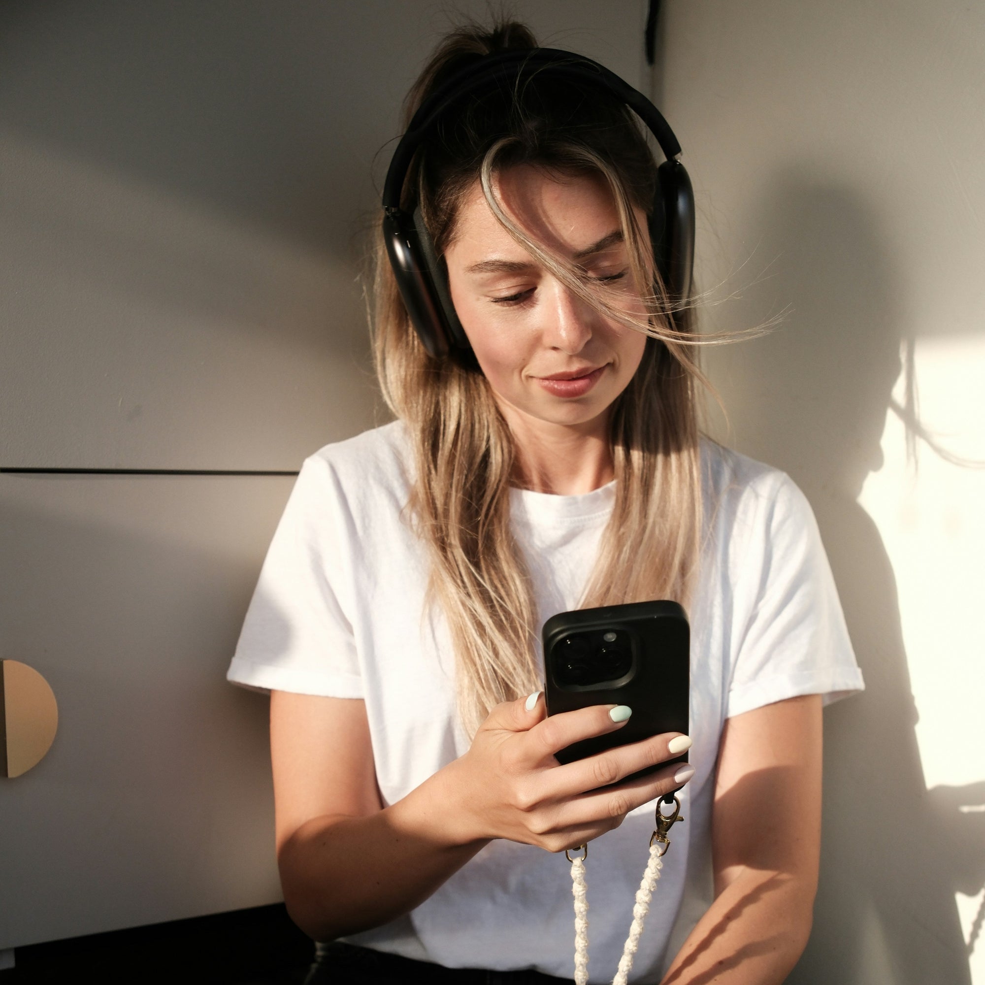 Eine Person sitzt im Sonnenlicht, trägt schwarze Kopfhörer und nutzt ein Smartphone, um einen Spiegel bei Spiegelmax online zu konfigurieren. Moderne, helle Umgebung, entspannte Atmosphäre.