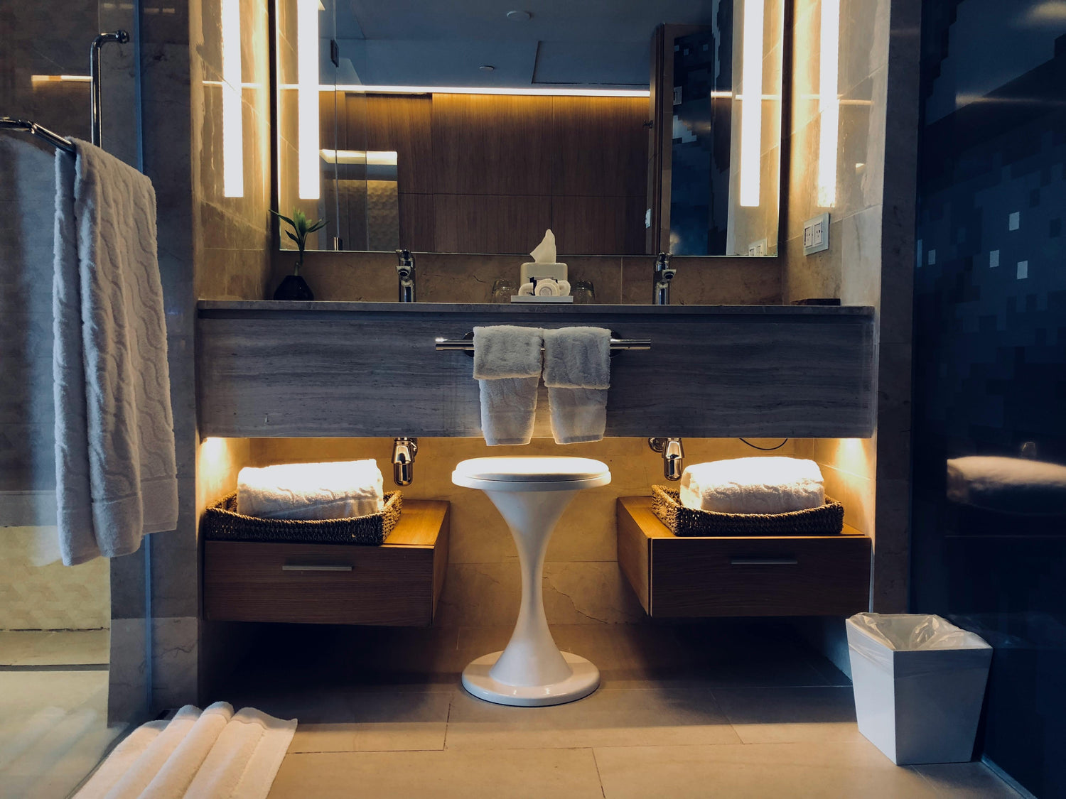 Modernes Hotelbadezimmer mit großem beleuchtetem Spiegel, zwei Waschbecken, Handtüchern auf Ablagekörben unter dem Waschtisch, warmen Lichtakzenten und hellem Hocker in der Mitte.