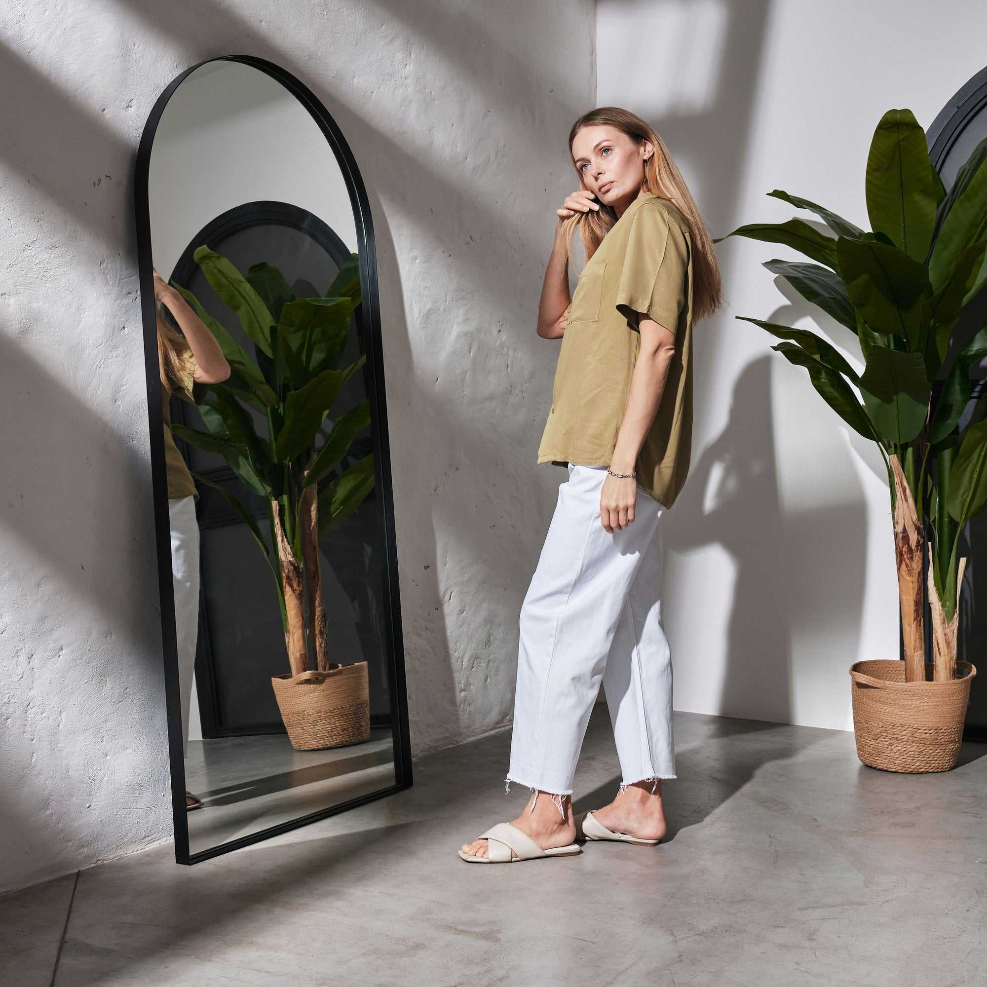 Moderner, hoher Standspiegel mit gebogenem Rahmen von Spiegelmax, vor dem eine Frau in heller Kleidung steht. Der Spiegel reflektiert eine große Zimmerpflanze, während Sonnenlicht und Schattenmuster den minimalistischen Raum akzentuieren.