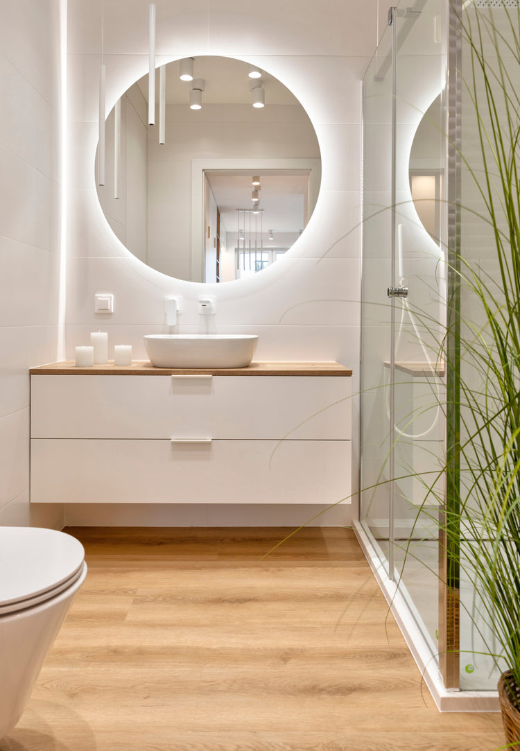 Ein modernes, hell gestaltetes Badezimmer mit einem großen runden LED-Spiegel über einem weißen Waschtisch mit Holzplatte. Auf dem Waschtisch steht ein weißes Aufsatzwaschbecken, daneben mehrere Kerzen. Rechts befindet sich eine Glasdusche, deren Türen den Spiegel reflektieren. Heller Holzfußboden und schlichte weiße Wände geben dem Raum ein warmes, minimalistisches Ambiente; im Vordergrund wächst eine dekorative Pflanze.