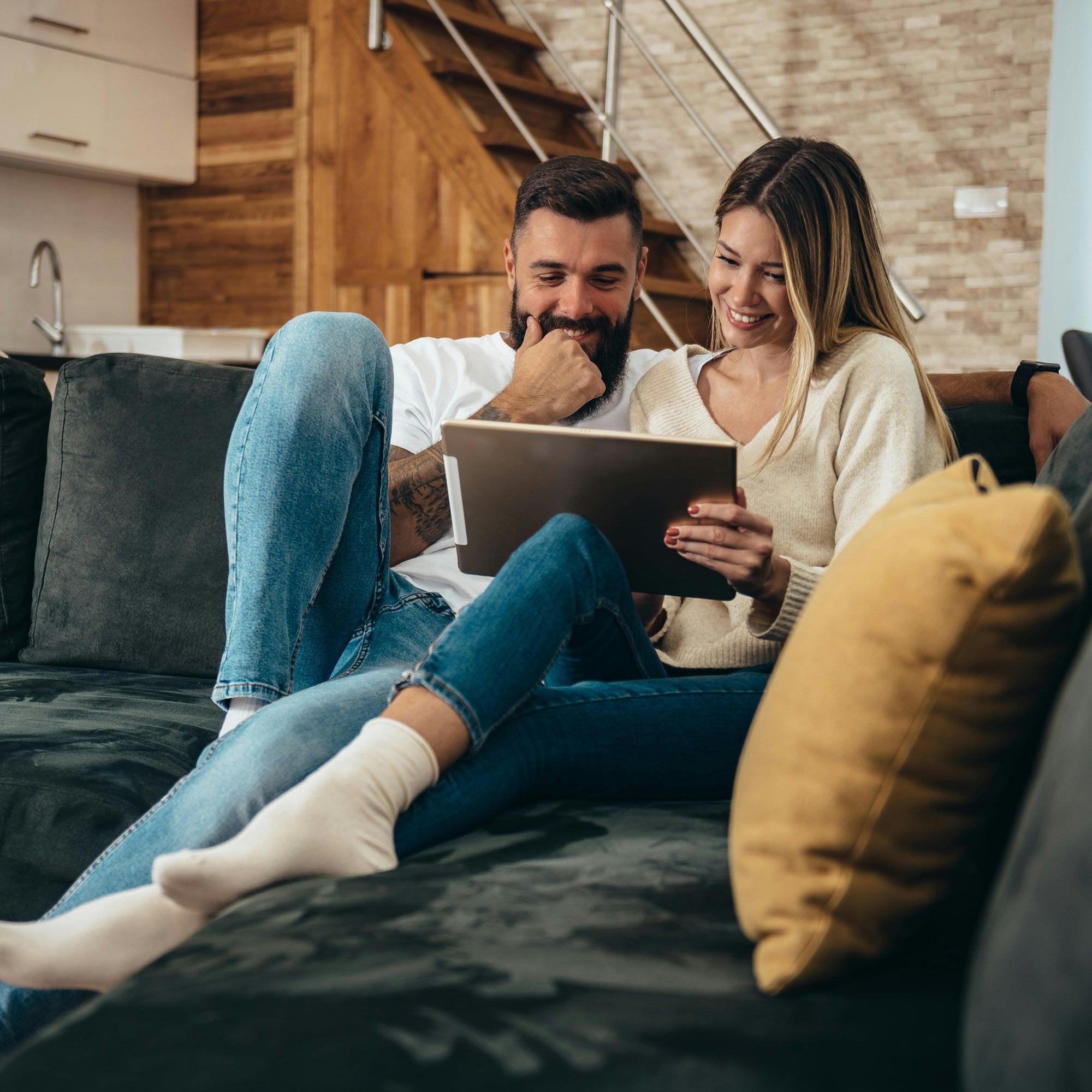 Ein Paar sitzt entspannt auf dem Sofa und bestellt über ein Tablet einen Spiegel von Spiegelmax. Der gemütliche Wohnbereich mit warmen Holzelementen bildet den Hintergrund.