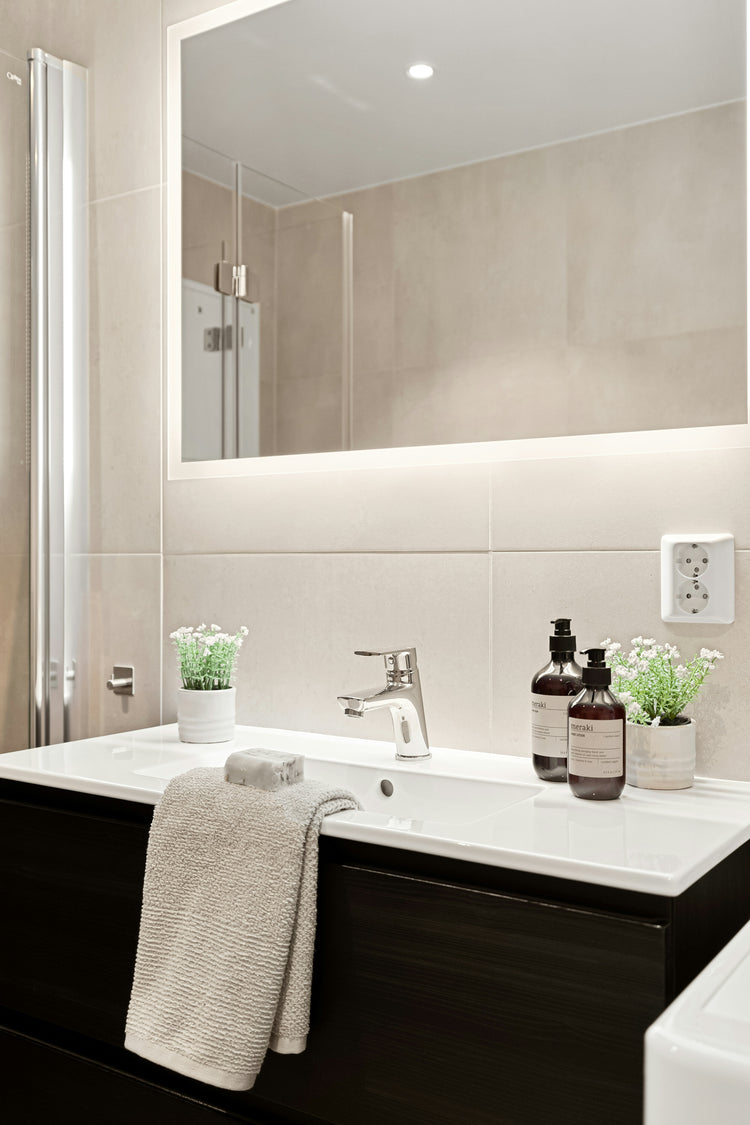 Ein moderner LED-Badspiegel von Spiegelmax mit umlaufender Beleuchtung hängt über einem weißen Waschtisch mit dunklem Unterschrank. Vor dem Spiegel stehen kleine Pflanzen und Pflegeprodukte, ein Handtuch liegt über dem Waschbeckenrand. Die hellen Fliesen und die klare LED-Beleuchtung des Spiegels sorgen für ein minimalistisches, freundliches Badezimmerambiente.