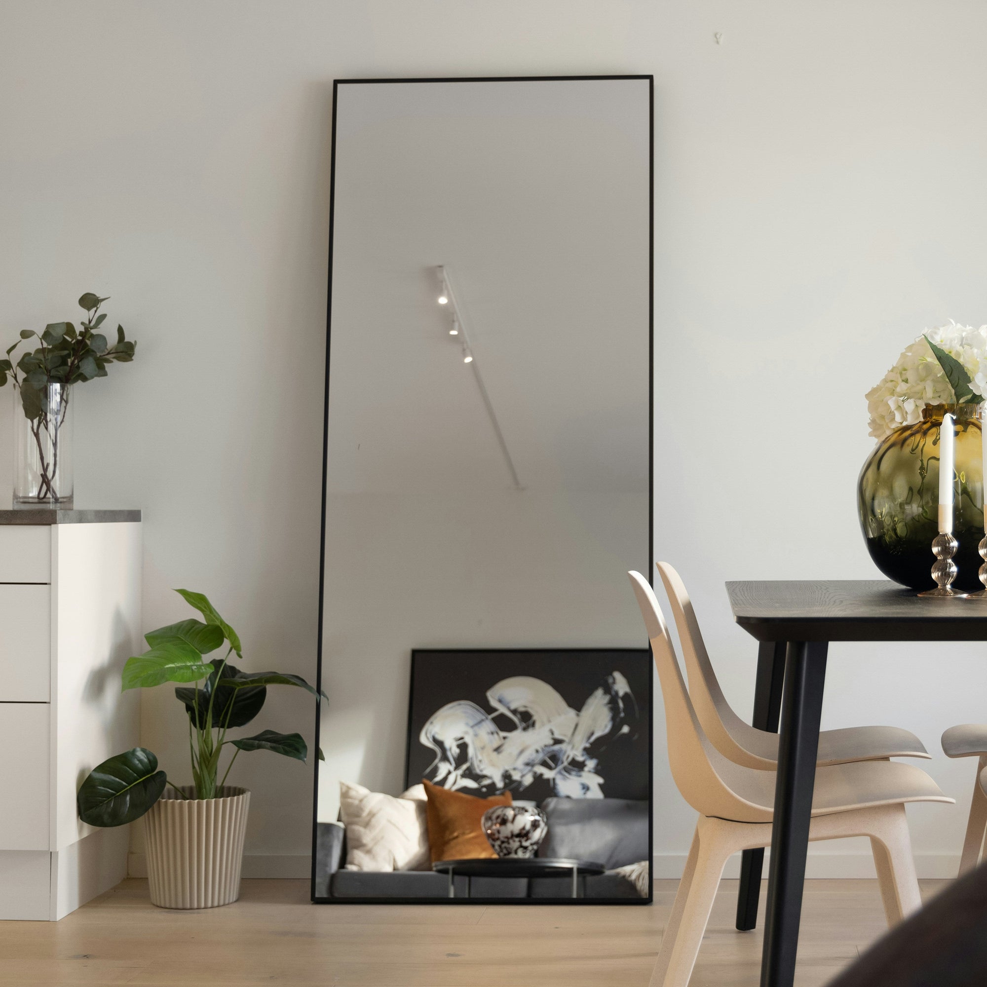 Großer rechteckiger Standspiegel von Spiegelmax mit schwarzem Rahmen, der ein stilvoll eingerichtetes Wohnzimmer mit Kunstbild, Sofa und Pflanzen reflektiert. Der Spiegel steht an einer weißen Wand neben Esstisch und Dekoration.