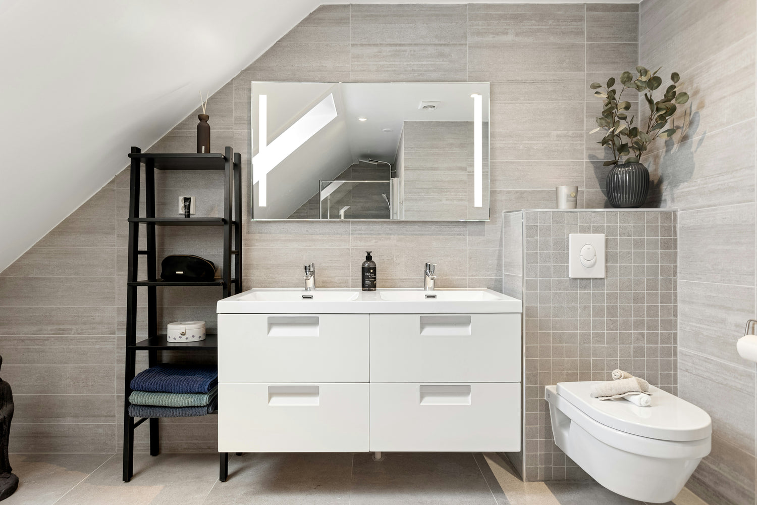 Modernes Badezimmer mit schräger Decke, weißem Doppelwaschbecken, großem beleuchtetem Spiegel, wandhängender Toilette und offenem schwarzen Regal mit Handtüchern und Accessoires, umgeben von hellgrauen Wand- und Bodenfliesen.