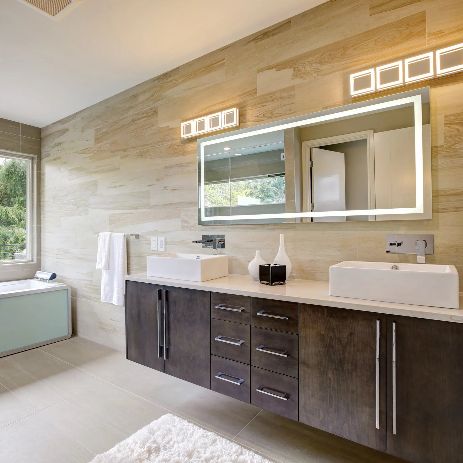 Geräumiges modernes Badezimmer mit hellem Naturstein an der Wand, großem LED-beleuchtetem Spiegel, zwei eckigen Aufsatzwaschbecken auf dunklem Holzmöbel, wandhängender Armatur und Badewanne am Fenster.
