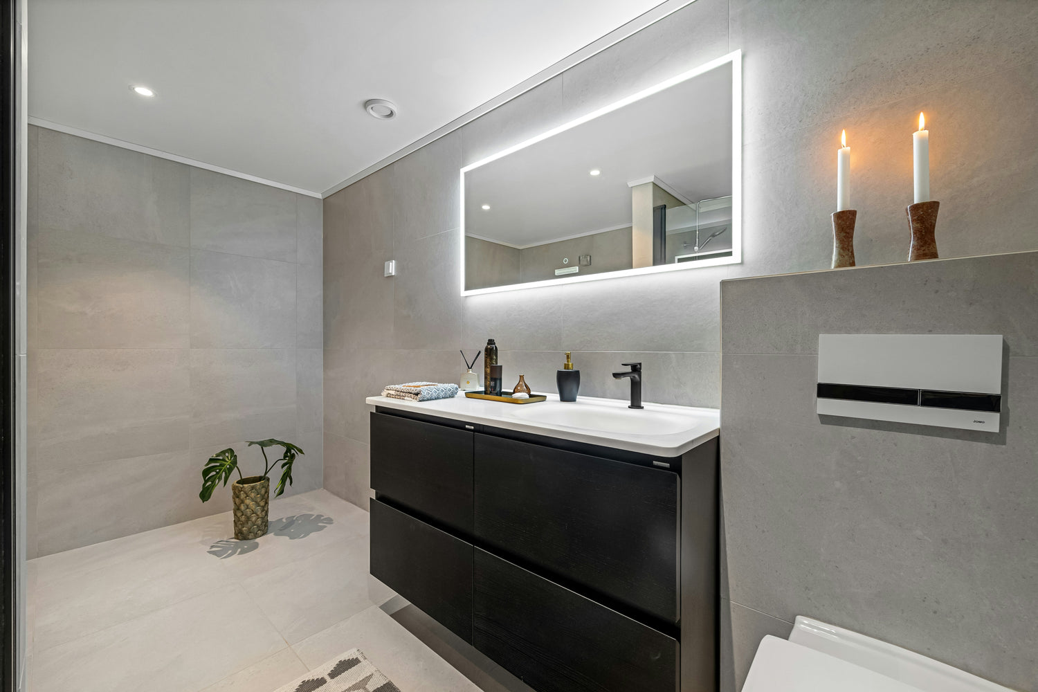 Modernes Badezimmer mit großem LED-beleuchtetem Spiegel, dunklem Waschtisch, schlichten grauen Fliesen, dekorativen Kerzen und einer grünen Topfpflanze auf dem Boden.