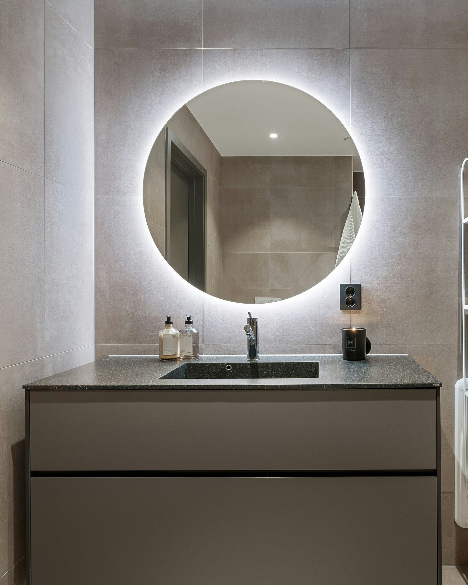 Modernes Badezimmer mit großem rundem LED-beleuchtetem Spiegel über einem grauen Waschtisch, schwarzer Armatur sowie dezenten Accessoires wie Seifenspendern und einer Kerze.