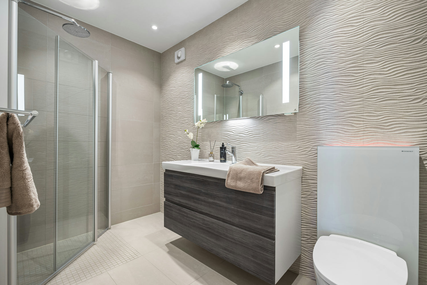 Modernes Badezimmer mit großer Glasdusche, strukturierter sandfarbener Akzentwand, beleuchtetem Spiegel, dunklem Waschtischunterschrank und wandhängender Toilette.