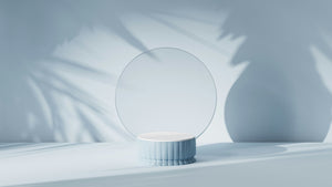 Runder, minimalistischer Schminkspiegel auf einem pastellblauen, geriffelten Sockel. Weiche Schatten von Pflanzen fallen auf den hellen Hintergrund und erzeugen eine ruhige, moderne Atmosphäre.