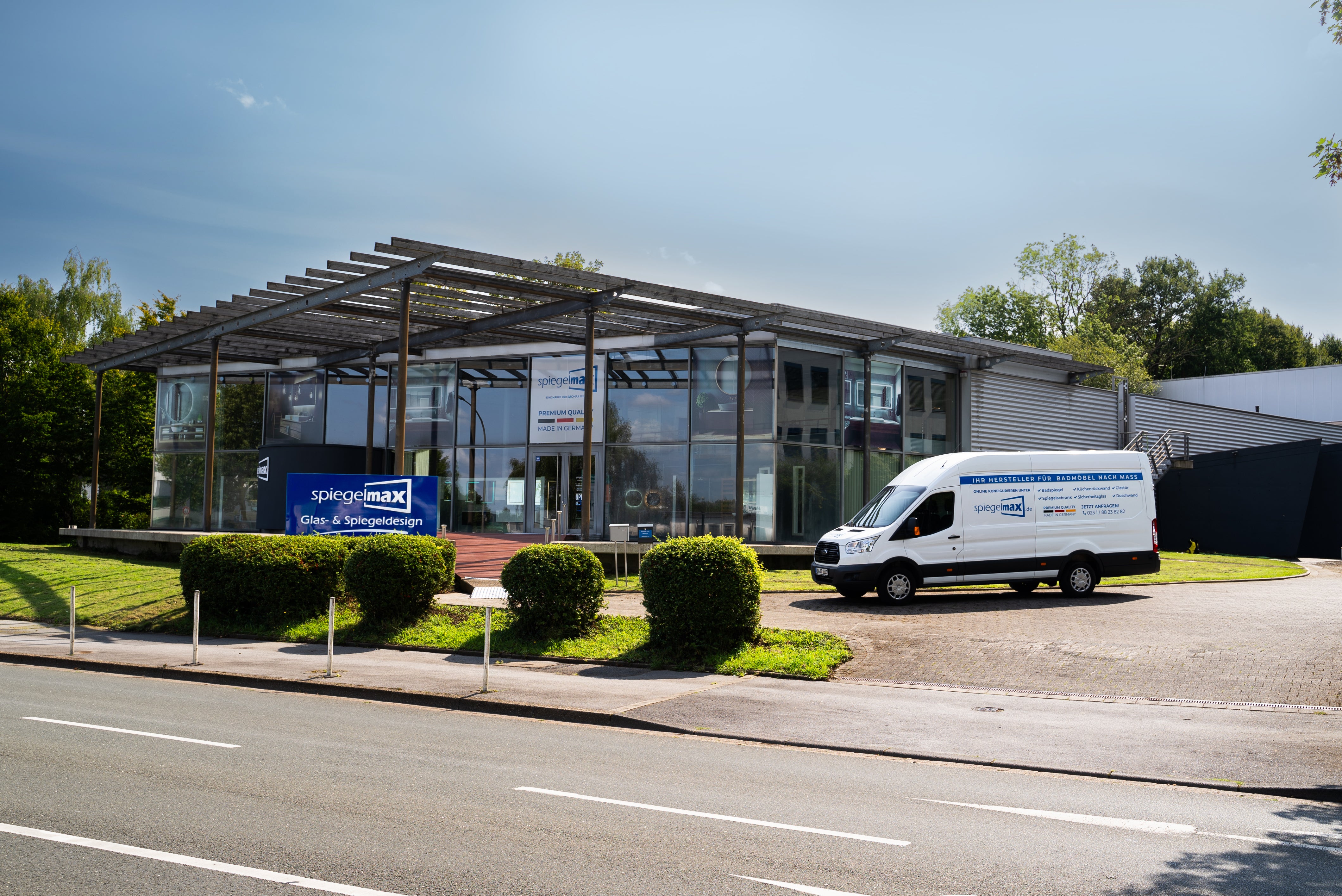 Das moderne Unternehmensgebäude von Spiegelmax mit großer Glasfassade, Showroom und sichtbarem Firmenschild „Glas- & Spiegeldesign“. Auf dem Vorplatz steht ein weißer Spiegelmax-Lieferwagen mit Markenlogo und Leistungsübersicht. Die Szene zeigt den Hauptsitz des Herstellers für LED-Spiegel, Badspiegel und maßgefertigte Spiegelprodukte in einer begrünten Umgebung.