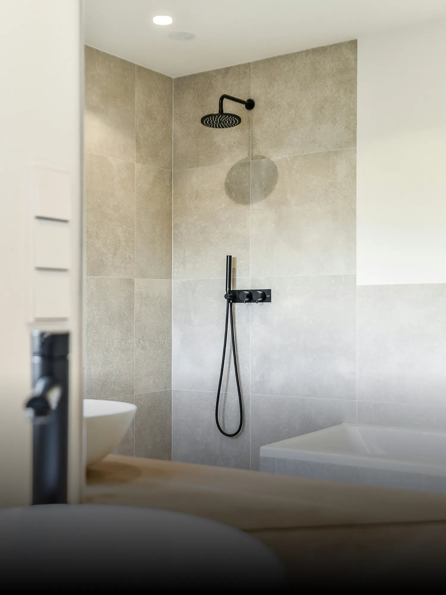 Dusche mit schwarzer Regendusche und Handbrause vor großformatigen Fliesen, ergänzt durch eine dezente Duschwand.