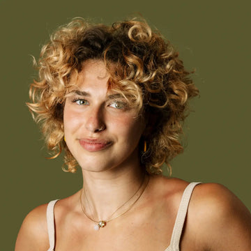 Porträt einer jungen Frau mit blonden, lockigen Haaren und grünlichen Augen vor olivgrünem Hintergrund. Sie trägt ein helles Top, eine feine Halskette und schaut freundlich in die Kamera.